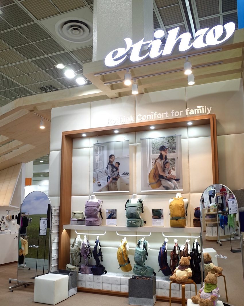 The Etihw stand at the fair, its lit sign above a wall of NUBE carriers in mint, lilac and mustard.