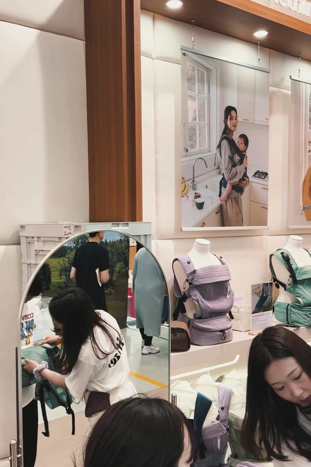 The Etihw stand at a Korean baby fair: NUBE carriers displayed on the wall in mint and lilac, a customer trying one on at the fitting mirror while staff assist.